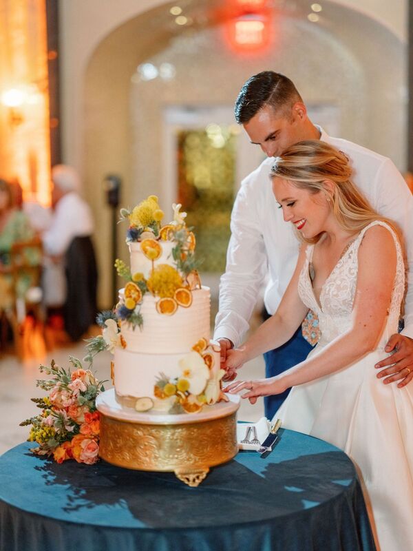 bride and groom cutting a three-tiered white wedding cake with bright tropical flowers and dried citrus decorations