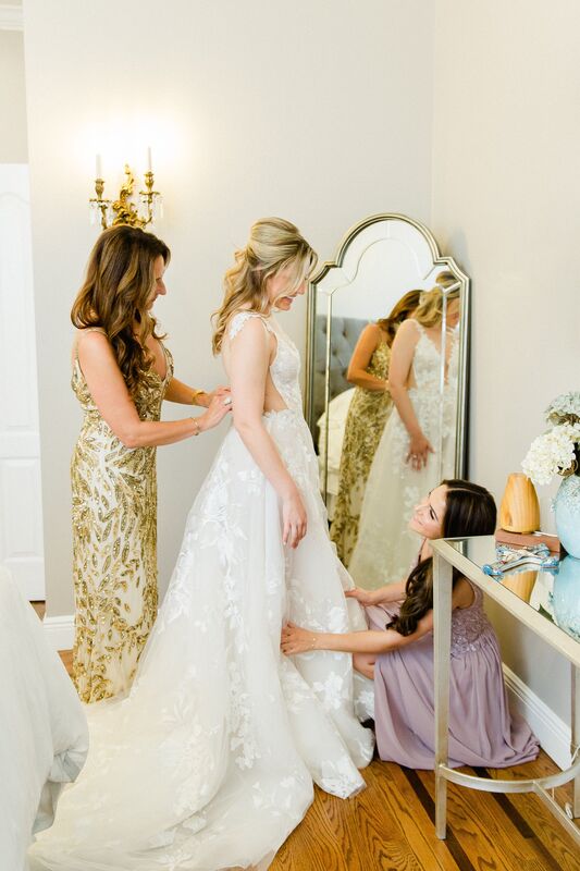 bride standing in front a n oval mirror as her wedding party helps her dress