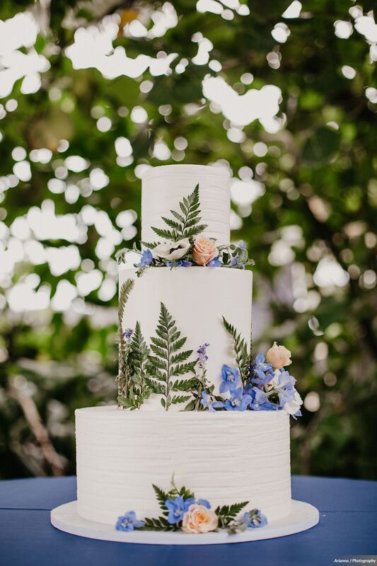 Three tiered white wedding cake with pressed ferns and pink and blue flowers